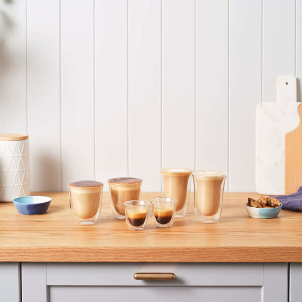 Six verres à double paroi transparents De’Longhi de différentes tailles sur un plan de travail en bois, avec espresso et cappuccino à l’intérieur