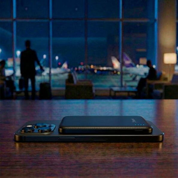 Mise en situation d'un smartphone Android avec batterie externe posée dessus sur une table en bois dans un grand hall d'un aéroport avec baie vitrée donnant sur les pistes d'atterrissage de nuit