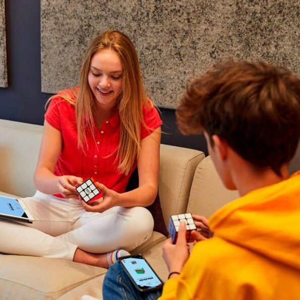Concours de speedcubing entre une jeune fille et un jeune garçon assis sur un canapé avec chacun un Rubik's Cube et un smartphone avec application compatible