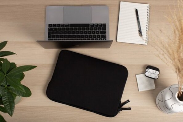 Housse de protection noire posée sur un bureau en bois clair à côté d'un MacBook, d'une plante verte, d'une herbe de pampa et d'un bloc notes avec crayon