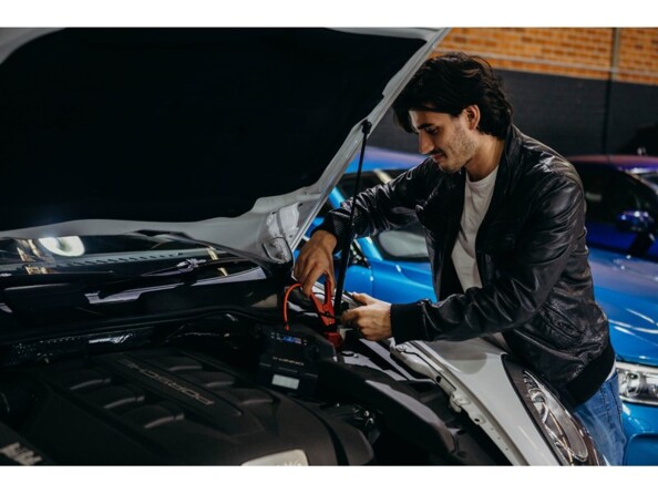 Jeune homme en veste en cuir entrain de brancher les pinces crocodiles du booster à la batterie de sa voiture avec le capot ouvert