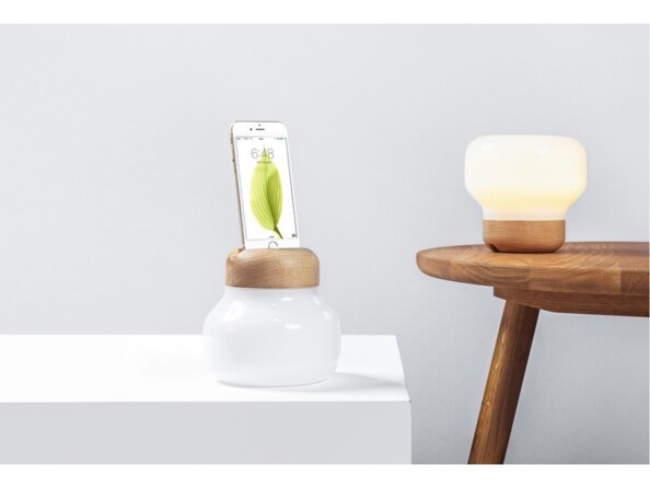 Lampe d'ambiance avec station de chargement Lightning en cours de recharge d'un iPhone posée sur un meuble blanc à côté d'un tabouret en bois sur lequel se trouve une seconde lampe LED du même type