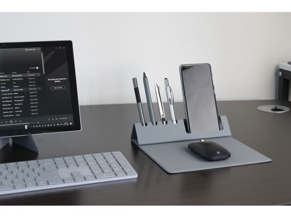 Mise en situation du pad de souris pliable avec support pour téléphone et quatre porte-stylos sur un bureau à côté d'un PC fixe avec clavier sans fil