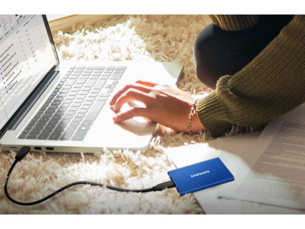 PC portable posé au sol sur un tapis devant une personne assise et branché par câble USB à un disque dur SSD T7 Samsung