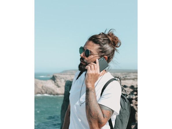 Homme barbu avec t-shirt blanc, chignon et lunettes de soleil entrain de téléphoner devant la mer avec un sac à dos sur le dos