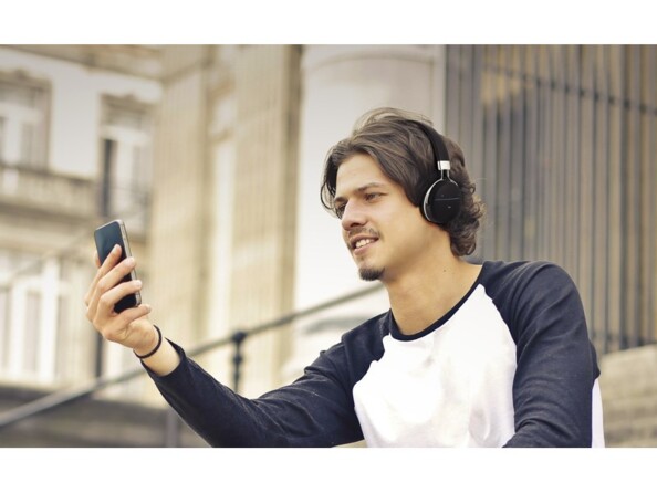 Homme trentenaire assis sur des escaliers extérieurs en appel vidéo avec son téléphone portable et le micro de son casque sans fil