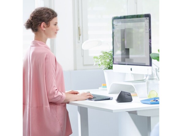 Mise en situation d'une jeune femme brune en blazer rose pâle entrain de travailler à son bureau debout sur ordinateur dans un setup informatique moderne