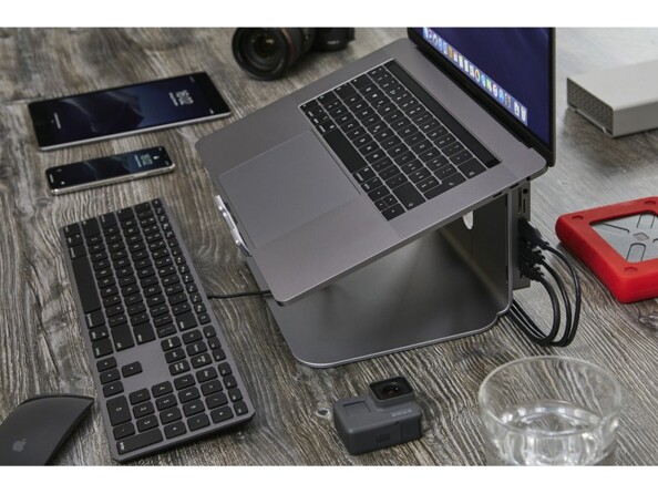 Setup de bureau avec MacBook surélevé sur support avec hub USB, clavier filaire, disque dur externe, tablette, smartphone et verre d'eau sur un bureau aspect bois
