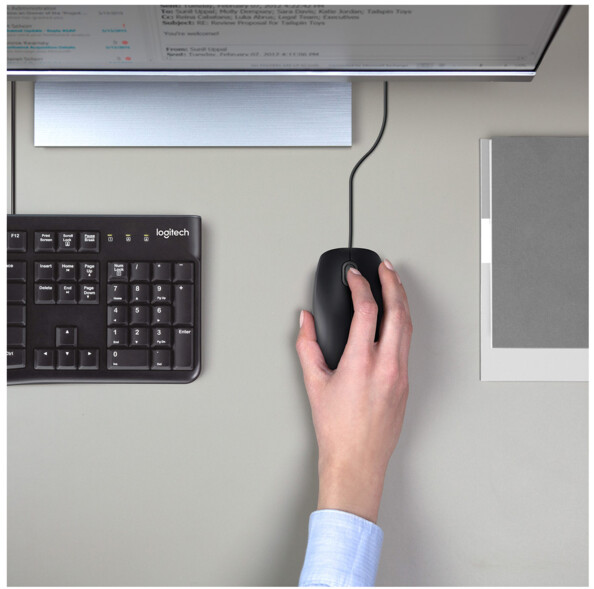Mise en situation de la souris optique filaire USB Logitech dans la main d'une personne en chemise sur un bureau à côté de documents, d'un clavier et d'un écran PC