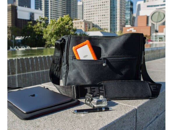 Mise en situation du HDD LaCie Rugged Mini rangé dans une poche d'un sac en bandoulière posé sur un muret en pierre en extérieure à côté d'un PC portable fermé et d'une Go Pro