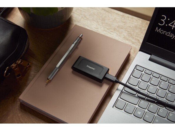 Mise en situation du SSD externe Kingston branché par câble USB à un PC portable sur un bureau en bois à côté de fournitures de bureau