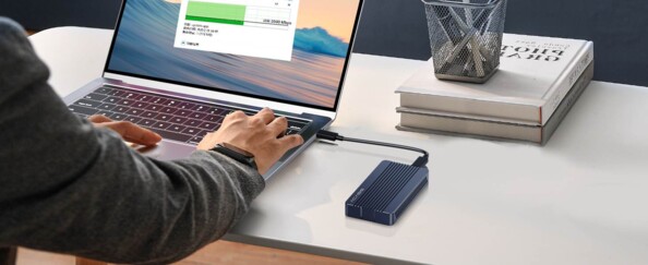 Mise en situation du boîtier pour SSD NVMe PCIe 2230/2242/2260/2280 coloris bleu branché au port USB-C d'un PC portable sur lequel travaille une personne assise à son bureau à l'aide du câble USB-C fourni en accessoire