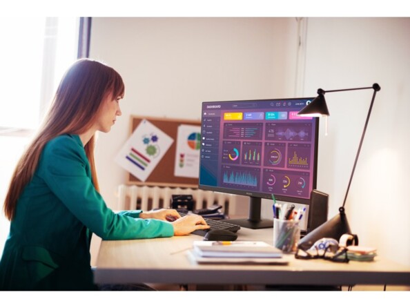 Jeune femme habillée d'un pull à manches longues verts les cheveux longs bruns avec franche assise devant un bureau et travailler sur PC fixe à côté d'un radiateur et de diverses fournitures de bureau