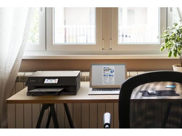 Mise en situation de l'imprimante jet d'encre 3 en 1 Canon coloris noir sur un bureau en bois type meuble scandinave devant une fenêtre à côté d'un PC portable allumé