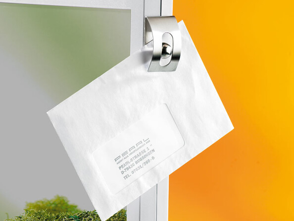 Mise en situation du porte-notes Infactory fixé à une surface lisse style cadre de porte avec enveloppe maintenu par bille en acier inoxydable