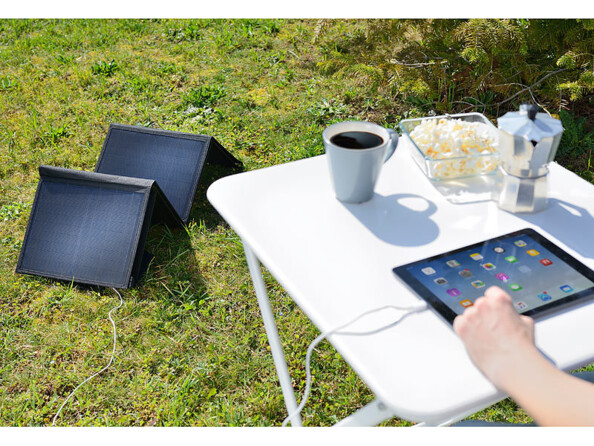 Mise en situation du module solaire plié sur le gazon d'un espace extérieur et branché à une tablette allumée en cours de chargement sur une table de camping blanche sur laquelle sont posés pop corn, cafetière et tasse de café