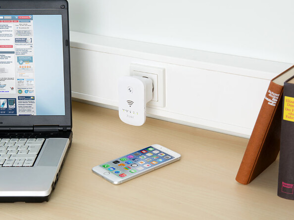 Mise en situation de l'amplificateur de réseau wifi 7Links branché à une prise secteur située au-dessus d'un plan de travail beige sur lequel sont posés des livres, un ordinateur et un iPhone allumés