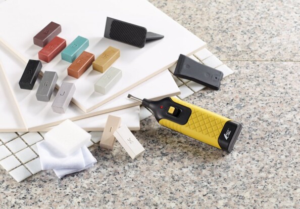 Fer à fondre posé sur le sol avec les différents outils présents dans la malette de rangement dont 11 bâtons de cire aux couleurs différentes à faire fondre avec le fer comme solution aux rayures, marques et cassures dans vos sols durs