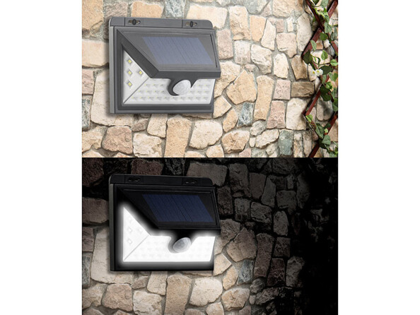 Mise en situation du spot LED solaire fixé sur un mur de pierre à côté d'une plante grimpante éteint de jour et allumé de nuit