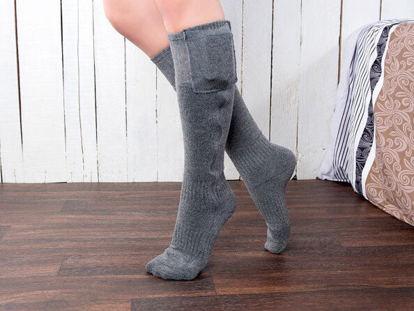 Personne portant aux pieds les deux chaussettes montantes et chauffantes sur un parquet en bois foncé dans une chambre aux murs en lambris blancs