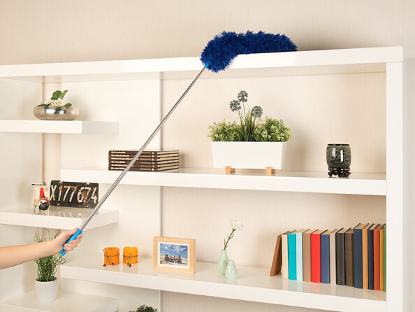 Main époussetant des étagères avec un plumeau télescopique bleu dans un intérieur avec livres et plantes