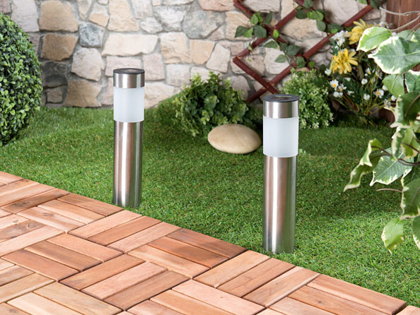 2 lampes solaires LED en acier inoxydable plantées dans le gazon, le long d'un chemin pavé d'un jardin, devant un mur en pierre sur lequel grimpe du lierre