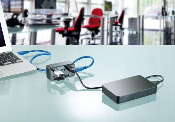 Mise en situation du hub USB branché à un disque dur externe et à un ordinateur portable via deux câbles USB sur un bureau en verre dans un open space