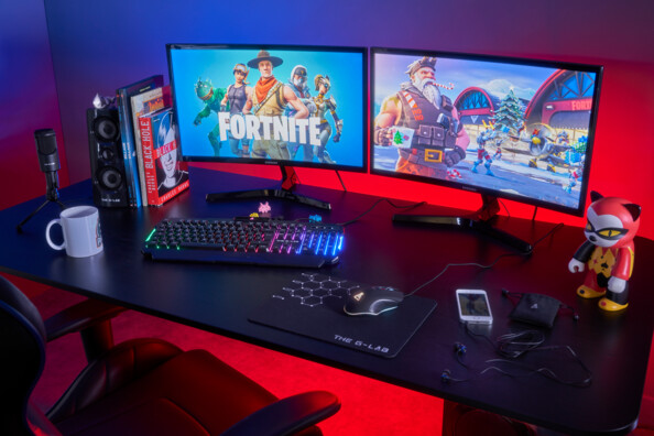 Setup gaming complet sur bureau avec pack gaming filaire combo Helium devant deux écrans d'ordinateurs avec jeu Fortnite, clavier et souris branchés aux écrans, pad sous la souris et écouteurs branchés à un smartphone