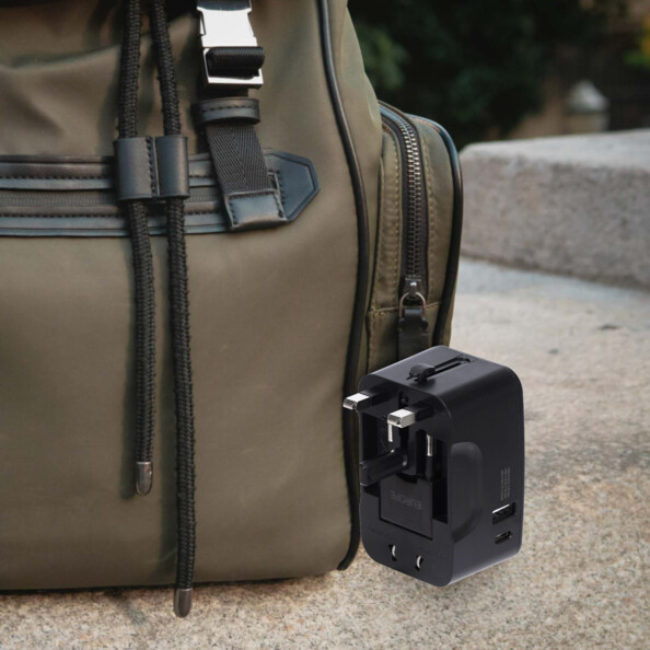 Adaptateur secteur de voyage noir posé sur une marche d'escalier à côté d'un sac à dos brun avec fiche UK déployée