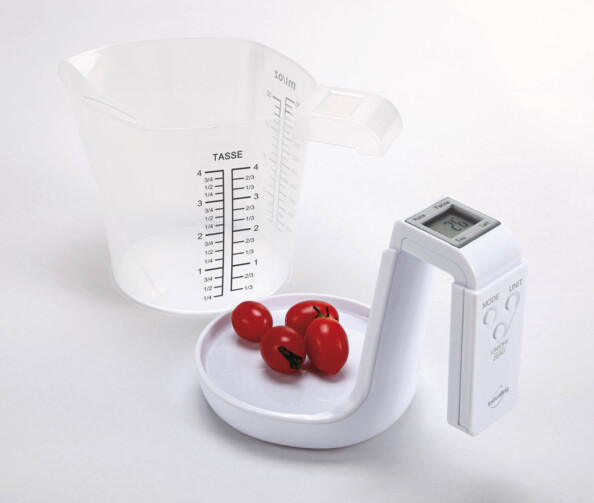 Pèse-aliments coloris blanc avec poignée dotée de boutons de commande et d'un écran LCD pesant des tomates cerises sur sa surface de pesée avec gobelet doseur à côté