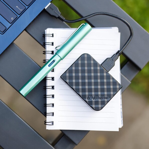 Mise en situation du disque SSD Verbatim 1 To design 3D noir branché à un ordinateur portable bleu métallisé et posé sur un bloc-notes avec un stylo bleu vert à côté