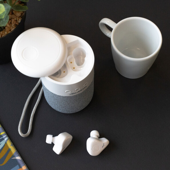 Vue du dessus de l'enceinte portable bluetooth avec boîtier de chargement des oreillettes sous le cache supérieur ouverte, dragonne en tissu attachée, écouteurs In-Ear sans fil sorti du boîtier et tasse vide posée à côté