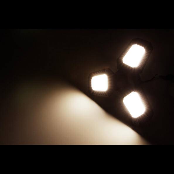 Mise en situation de la lampe LED fixée à un plafond d'une pièce sombre, éclairant la pièce d'une lumière blanc neutre par le biais de ses trois panneaux à LED
