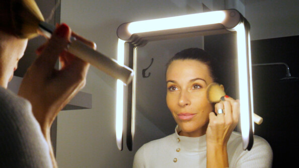 Femme souriante se maquillant les pommettes avec un gros pinceau devant le miroir avec la lampe pour miroir Wellys disposée en forme de pont sur le miroir par bande adhésive