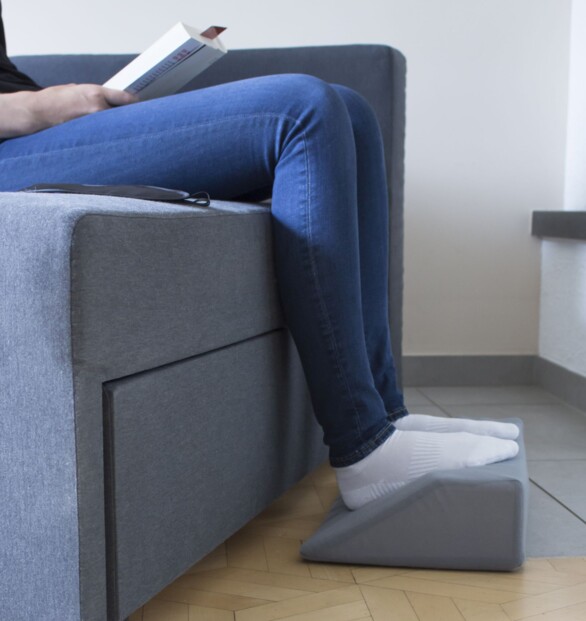 Personne assise entrain de lire un livre sur un canapé et gris, les pieds posés sur l'oreiller massant et chauffant lui-même posé sur le sol