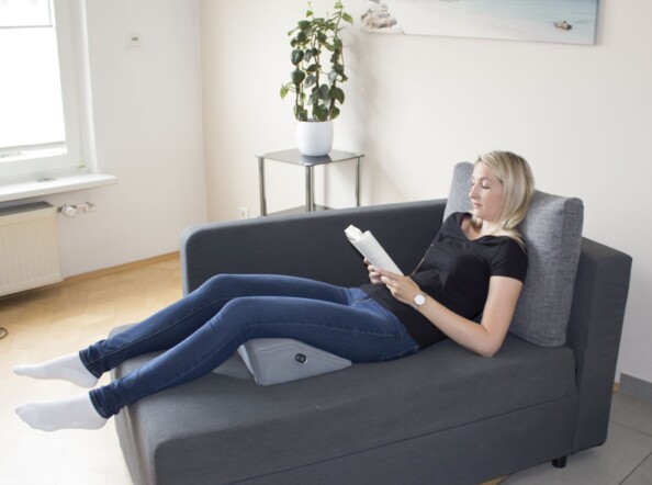 Jeune femme allongée sur un divan avec les cuisses surélevées par l'oreiller trapézoïdal chauffant, vibrant et massant