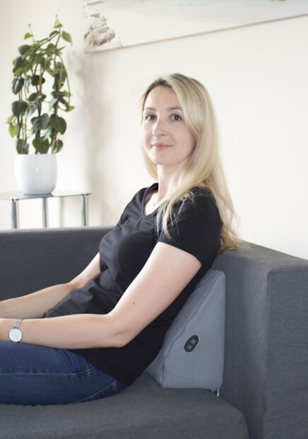 Femme blonde en t-shirt noir et jean assise contre l'accoudoir d'un canapé en tissu gris avec l'oreiller LH-001 Hydas entre son dos et le canapé, avec une plante grimpante dans un pot blanc en arrière-plan