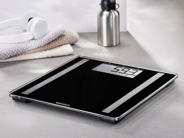 Balance moderne coloris noir avec 2 bandes de mesure du pourcentage en graisse et en eau contenu dans le corps, posée à côté d'une gourde, d'un casque audio et d'une serviette de sport