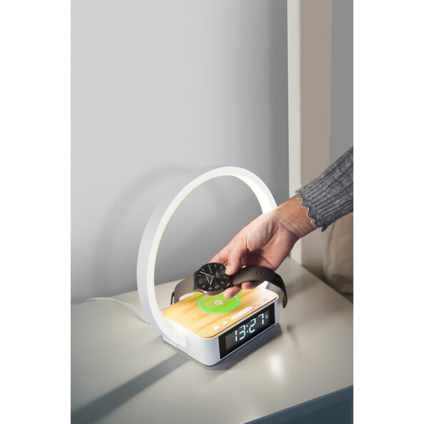 Mise en situation d'un homme en chemise posant sa montre connectée sur le chargeur à induction Qi de la lampe-réveil Hollywood placée sur sa table de chevet blanc brillant pour la recharger