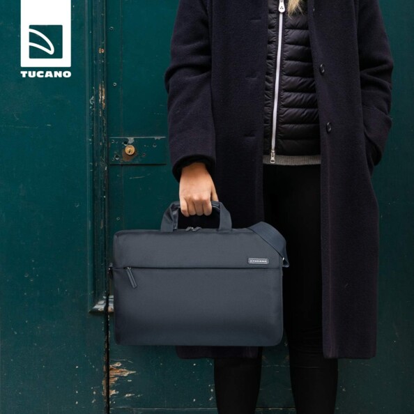 Femme en veste longue noir à boutons devant une porte d'entrée vert canard d'un immeuble tenant à la main la sacoche de protection pour PC portable et MacBook