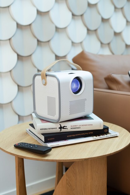 Vidéoprojecteur DV152 posé sur une pile de livres, sur une table en bois rond avec en arrière-plan un mur avec écailles vintage et un canapé en cuir marron