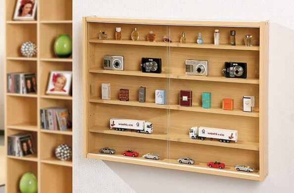 Mise en situation de la vitrine fixée sur un mur en papier peint blanc cassé, à côté d'une étagère au coloris similaire, dans laquelle sont rangés plusieurs types de produits : appareils photo, véhicules miniatures et échantillons de parfums