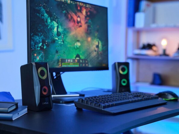 Mise en situation d'un setup gaming éclairé par une lumière bleue avec écran affichant une partie de LOL avec haut-parleurs Trust Gaming LED RVB branchés de chaque côté d'un clavier filaire