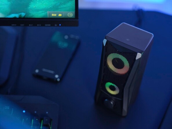 Vue du dessus d'une enceinte stéréo colonne avec rétroéclairage RVB, touche tactile et bouton de réglage du volume à côté d'un PC gamer et d'un smartphone affichant l'écran d'accueil