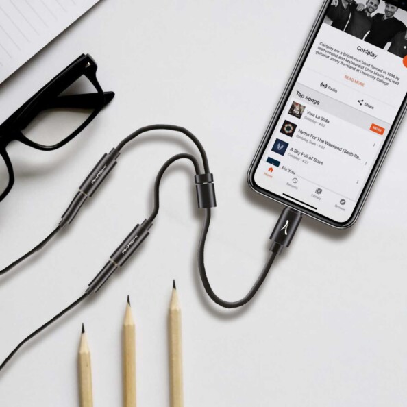 Câble adaptateur Lightning vers double jack branché à un iPhone affichant une playlist musical posé à côté d'une paire de lunettes et de trois crayons