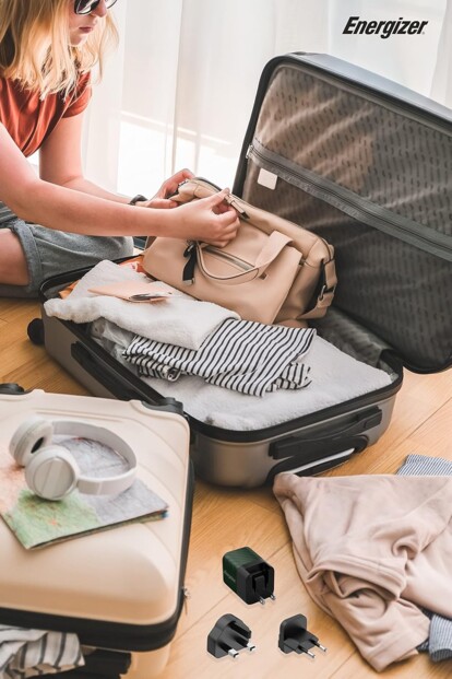 Vue du dessus d'une valise ouverte et en cours de remplissage d'effets personnels par une jeune femme avec adaptateur secteur de voyage EU/UK/US vers USB 20 W Energizer posé à côté