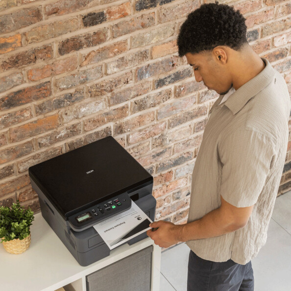 Jeune homme métisse entrain de récupérer une feuille imprimée en noir et blanc au format A4 sortant de son imprimante de bureau