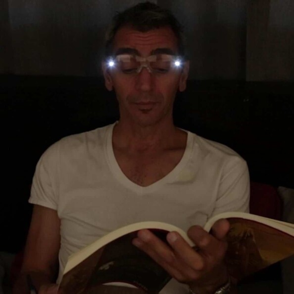 Homme en t-shirt blanc entrain de lire un bouquin dans le noir avec les lampes LED des lunettes allumées