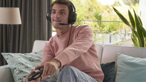 Jeune homme jouant en immersion à un jeu vidéo assis le canapé d'un salon avec manette en main et micro-casque Korp Cobalt sur les oreilles