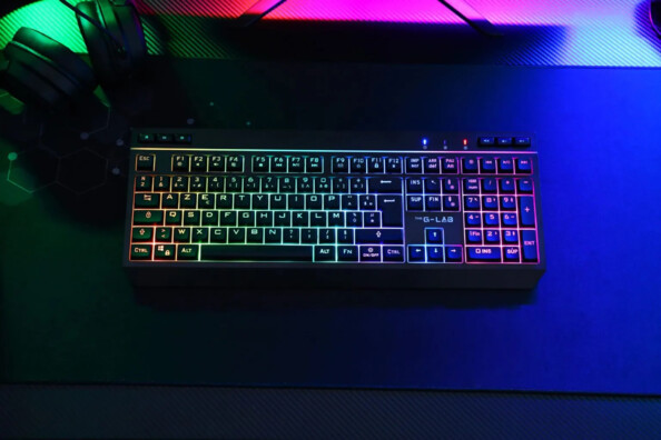 Clavier AZERTY LED RVB rétroéclairé personnalisable posé sur un tapis de bureau à côté d'un micro-casque filalire et d'un écran PC